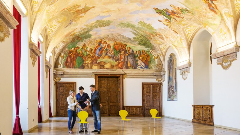 Interior view of a magnificent room in G&ouml;ttweig Abbey with frescoes on the ceiling and three people talking.