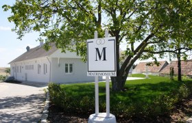 A white building with a "Matrimonium" sign in the foreground, surrounded by a green landscape.