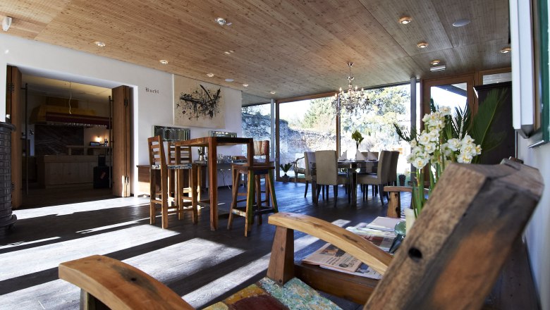 A modern breakfast room with wooden furniture, large windows and a chandelier. A wooden chair in the foreground and a dining table in the background.