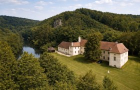 Aerial view of a large, historic building with a red roof, surrounded by forest and a river.