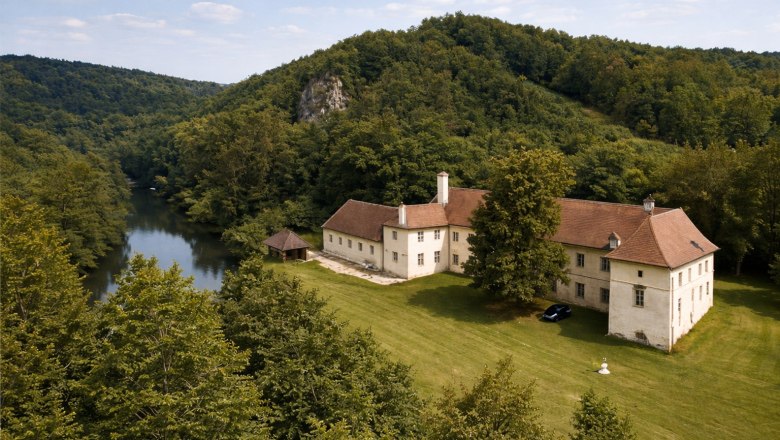 Aerial view of a large, historic building with a red roof, surrounded by forest and a river.