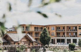 A modern wooden building with balconies and a traditional wooden house in the foreground, surrounded by trees and gardens.