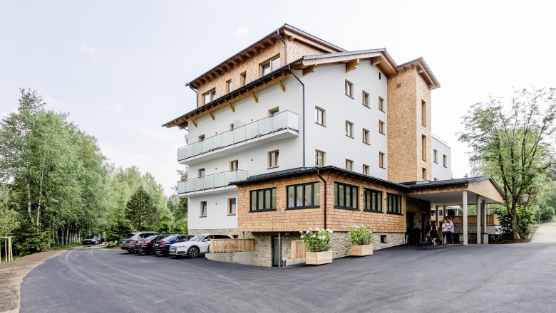A modern hotel building with wooden cladding and balconies, surrounded by trees and a parking lot with cars.