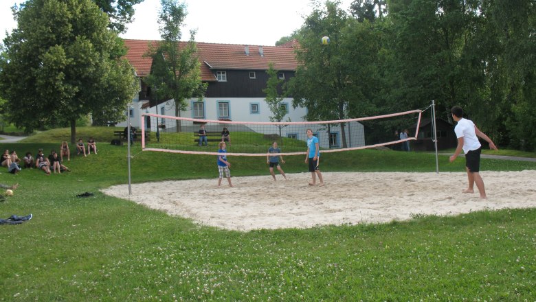 People playing volleyball on an outdoor sand court, surrounded by trees and a building in the background.
