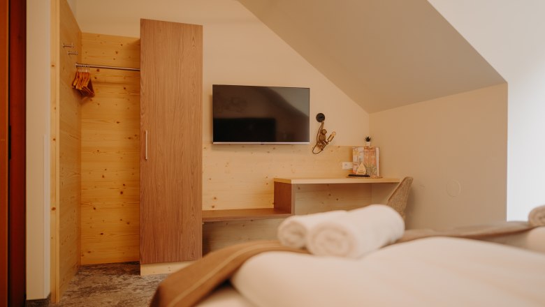 A cozy hotel room photographed from the bed. A flat-screen TV hangs on the wall across from the bed. Next to it is a light-wood wardrobe; part of the wall is also paneled with wood. There is also a shelf and a desk with a chair and a lamp. The room has a sloped ceiling.