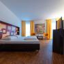 Modern hotel room with double bed, colorful wall pattern, two orange chairs and yellow curtains.
