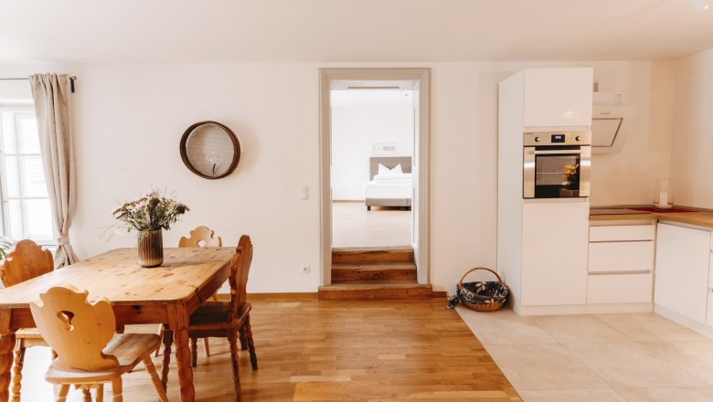 Bright kitchen with wooden table, chairs and modern white cupboards. Passageway to a bedroom in the background.