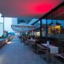 Terrace of a restaurant with tables and chairs, illuminated by red lights, with a view of buildings in the background.