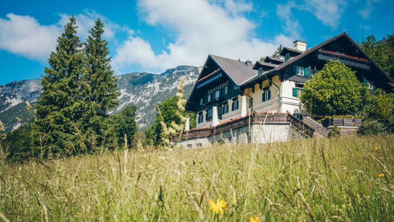 A traditional building called Knappenhof in a green meadow against a mountain backdrop.