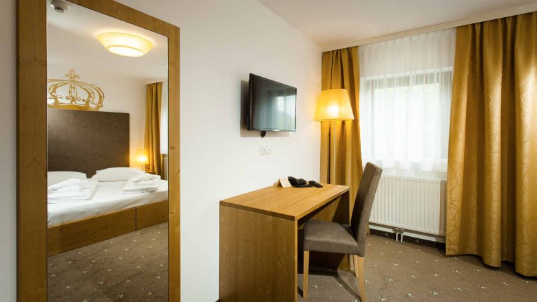A hotel room with a double bed, a desk and chair, a TV on the wall and yellow curtains.
