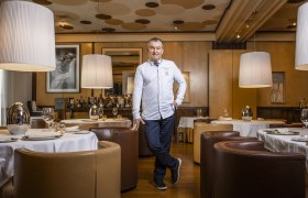 A man in a white chef's jacket stands in an elegant restaurant with round tables and large lamps.
