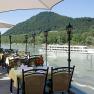Terrace with covered tables, view of the river and passing ship, surrounded by green hills.