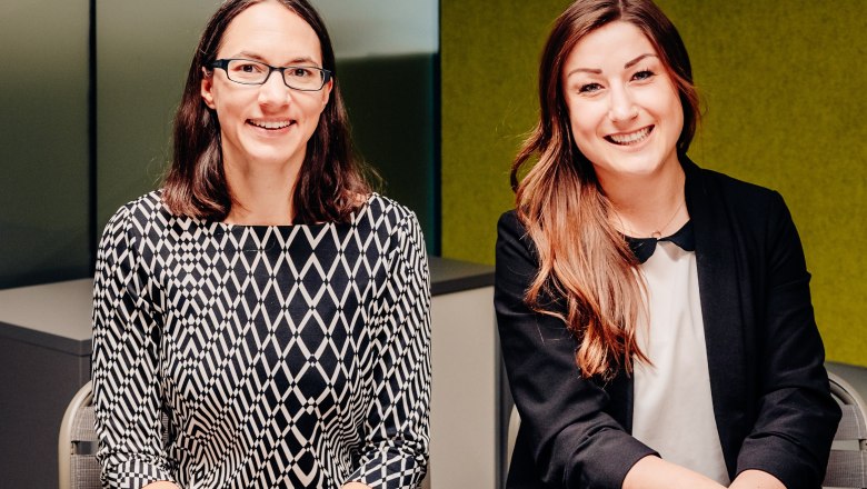 Two women sit next to each other and smile into the camera.
