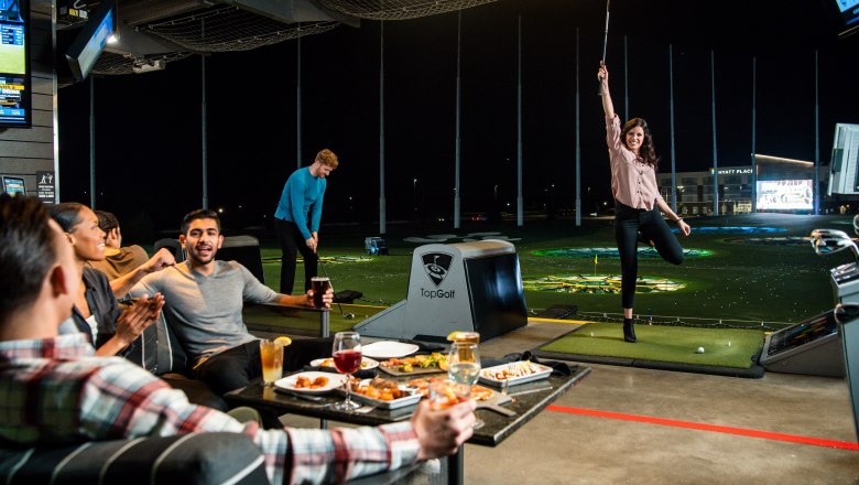 People play golf at TopGolf, a woman cheers while others eat and drink.
