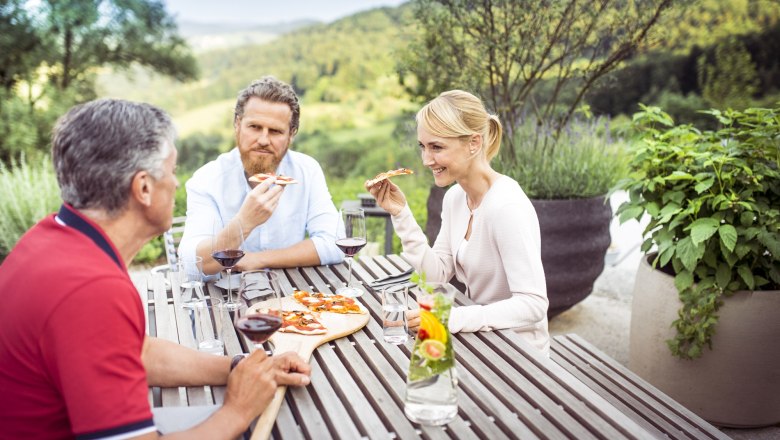 Three people eat pizza and drink wine at an outdoor wooden table overlooking a green landscape.