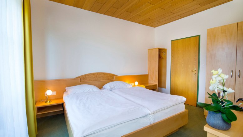 A simply furnished hotel room with a double bed, wooden ceiling, wardrobe and bedside lamps.