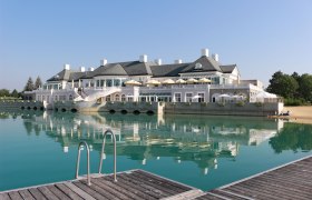 An elegant clubhouse on the lake with a terrace and sunshades, surrounded by clear water and a jetty in the foreground.