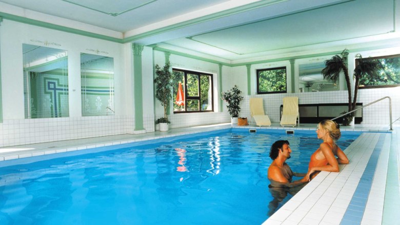 A couple in the pool of an elegant indoor area with plants and sun loungers.