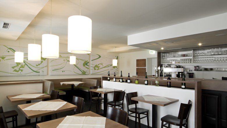 Interior view of a modern restaurant with wooden tables, chairs and a bar with bottles and glasses.