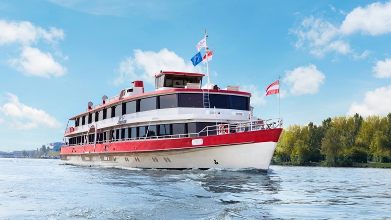 A passenger ship with red and white colors sails on a river under a blue sky.