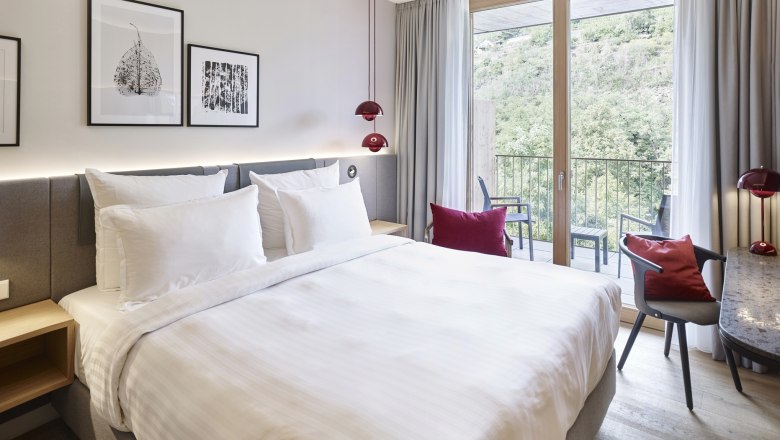 Modern hotel room with large bed, desk and balcony with a view of the green landscape.