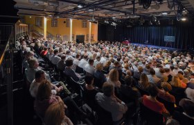 A packed theater hall with an audience looking onto a stage.