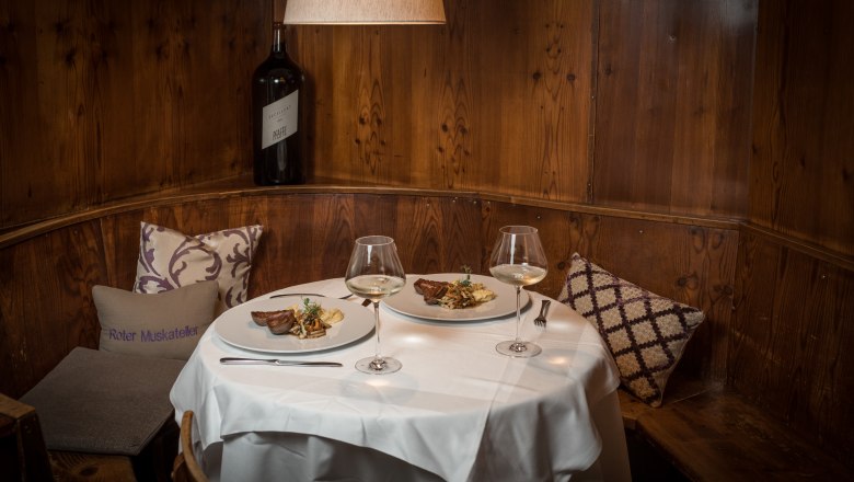 Cozy dining room with set table, two plates with food and wine glasses, wooden wall and cushions.