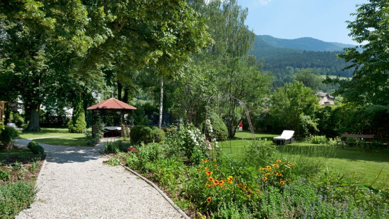 A well-kept garden with a gravel path, flower beds, a pavilion and sun loungers against a mountain backdrop.