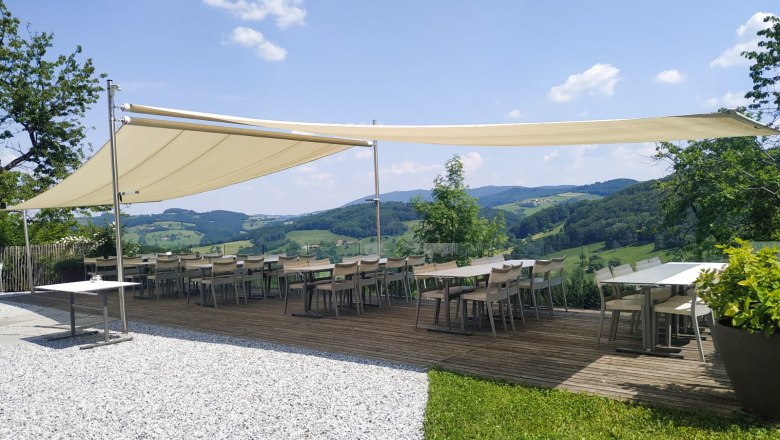 Terrace with awning and tables, surrounded by green landscape and blue sky.