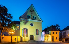 Yellow building with cross symbol, illuminated at night.