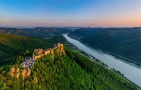 Aerial view of the ruins of Aggstein Castle on the Danube, surrounded by green hills and forests at sunset.