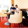 A couple is sitting on a bed in a hotel room, looking into a book. The room has red carpets and yellow curtains.