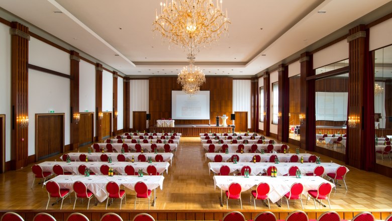 A large, elegant conference room with red chairs, white tablecloths and chandeliers.