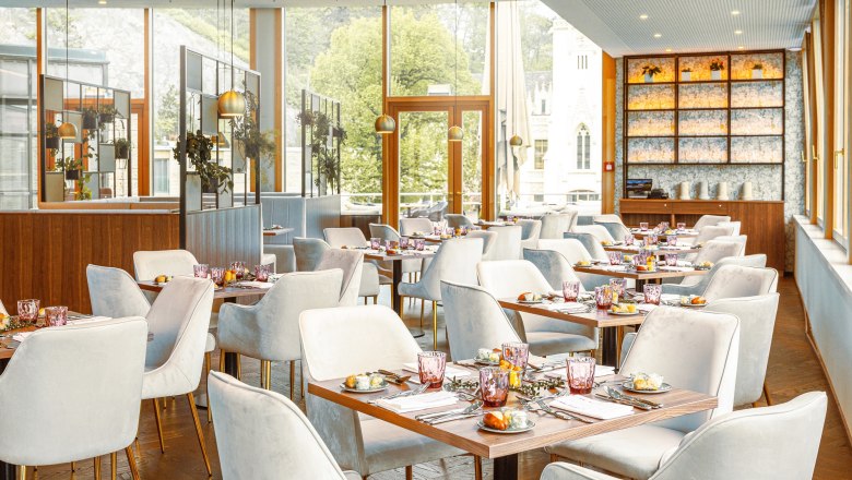 Elegant restaurant with laid tables and white chairs, large windows with a view of the greenery.