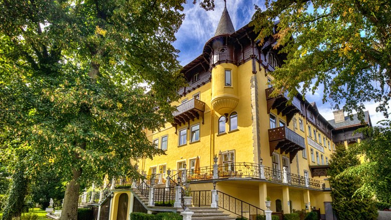 Historic yellow building with towers and decorations, surrounded by trees.