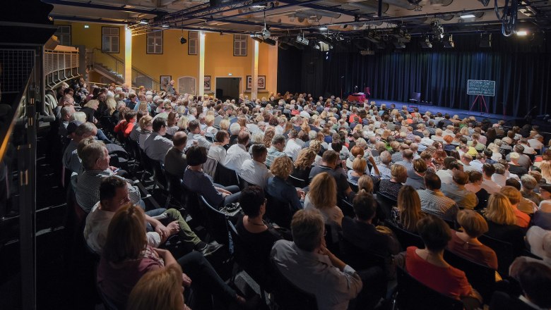 A packed theater hall with an audience looking onto a stage.