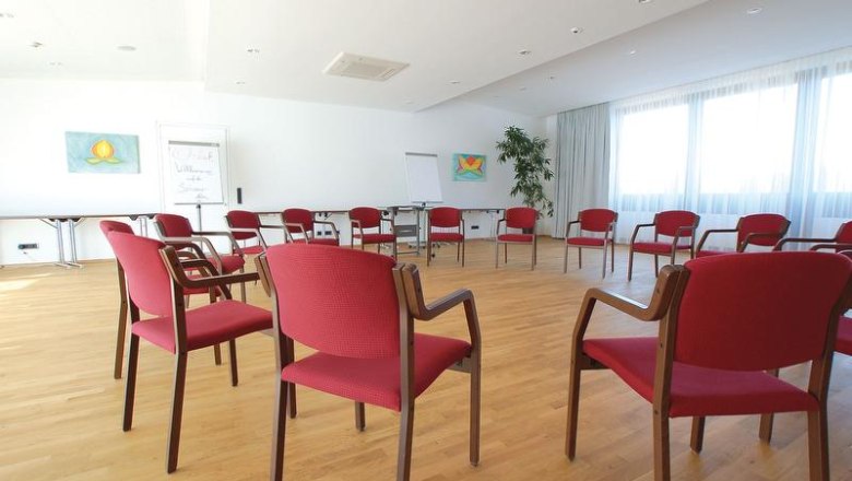 A bright seminar room with red chairs in a circle, a wooden floor and large windows.