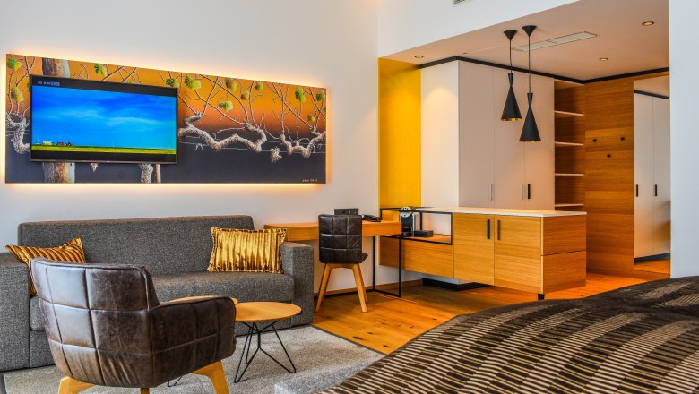 Modern hotel suite with sofa, armchair, TV and kitchenette.