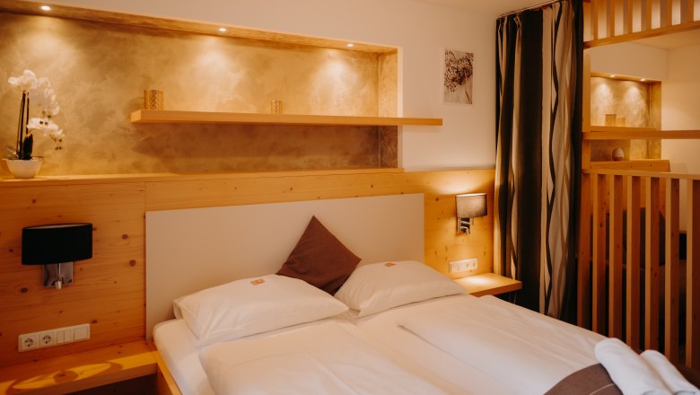 A cozy hotel room with a double bed, light-colored wood paneling, a wooden room divider, and a curtain. There are electrical outlets and light switches next to the bed. Above the bed is a wall niche that is illuminated and painted in an earthy gold tone.