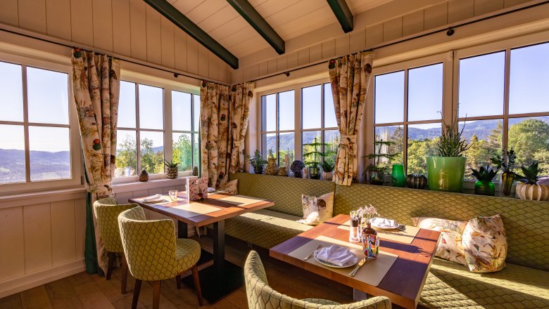 Cozy dining area with large windows, green upholstered furniture and plants, with a view of the countryside.