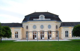 Historic building with symmetrical façade and large windows, surrounded by green spaces.