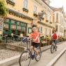 Two people are riding bicycles in front of a yellow hotel-restaurant with the sign 'Zur Post'.