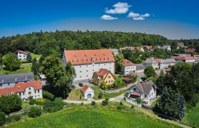 Aerial view of a village with a large white building with a red roof, surrounded by smaller houses and lots of greenery.