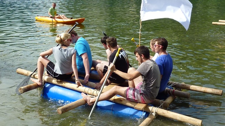 Group of people on a self-built raft on a lake, with a white flag and a kayaker in the background.