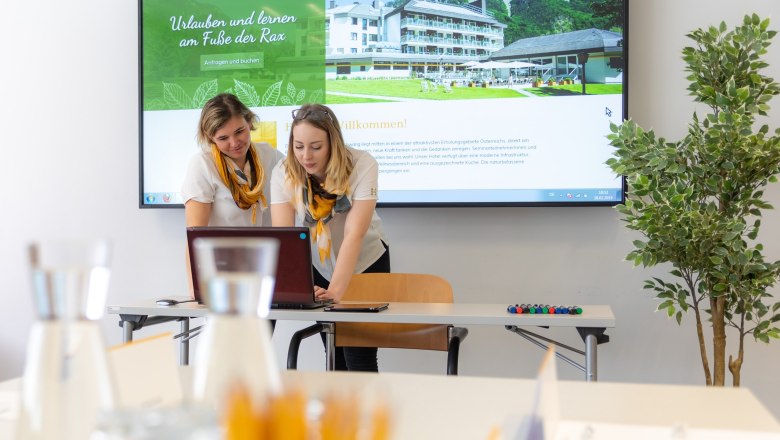 Two women work on a laptop in front of a large screen with a hotel website. Glasses and pens can be seen in the foreground.