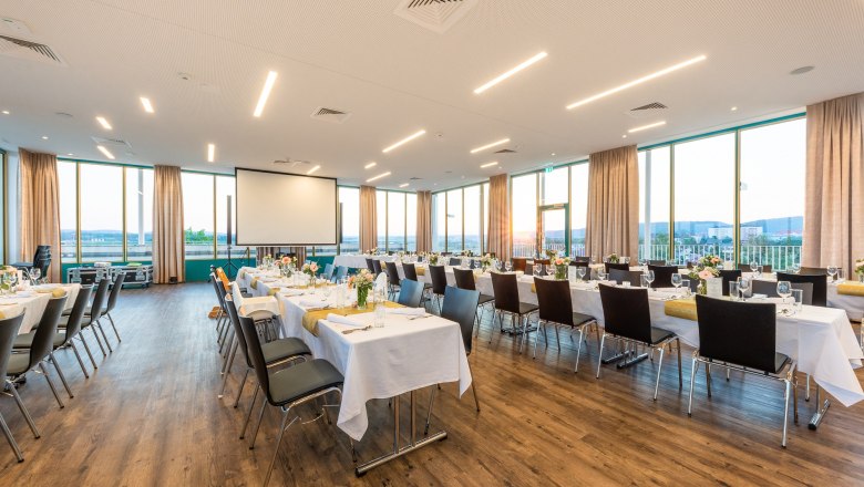 A large, bright room with covered tables and chairs, large windows and a projector.
