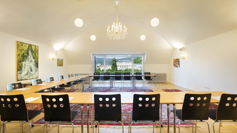 A conference room with a U-shaped table arrangement, chairs, a chandelier and large windows with a view of a building and mountains.