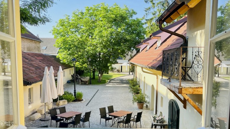 View from a window onto an inner courtyard with tables, chairs and parasols, surrounded by buildings and trees.