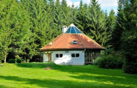 A round, white house with a pointed roof in the middle of a green forest.