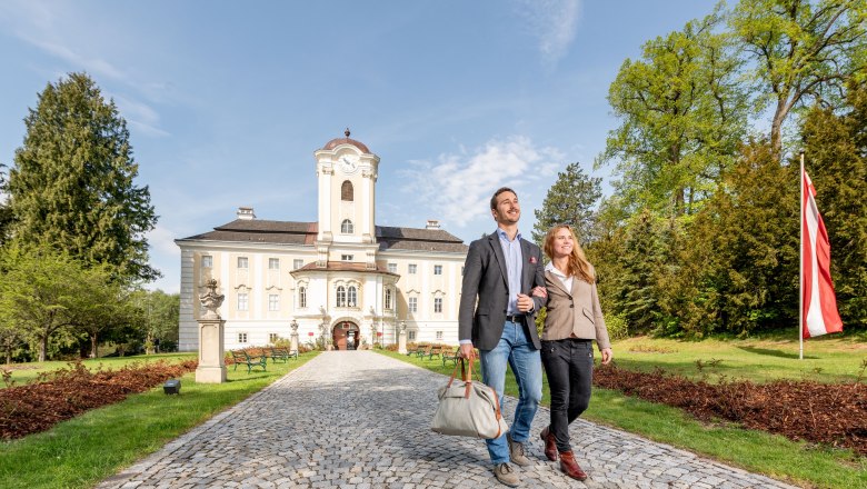A couple walks along a cobbled path in front of the Schlosshotel Rosenau, surrounded by green trees and an Austrian flag.